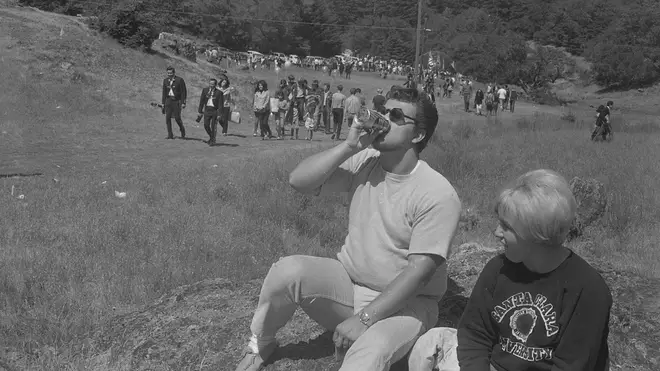 Man chugs beer at festival; partner looks on disapprovingly. A scene to be repeated at rock festivals for the next half century and beyond. Sir, we salute you.