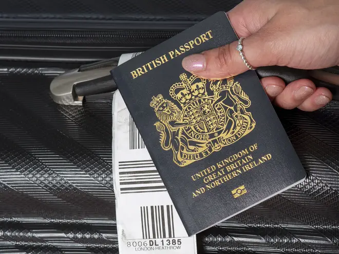 Dark blue colored British passport held in a travellers hand with holiday suitcase.
